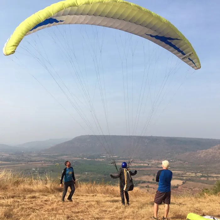 Guides helping a pilot launch a yellow wing from a dry grassy hilltop above a wide valley